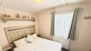 A bedroom with a wooden headboard, a bed with white bedding, a bedside table, a shelf with decorative items, a window with curtains, and a radiator. - Golden Palm The Retreat 38, Sleeps 6 Pet Friendly (Skegness)