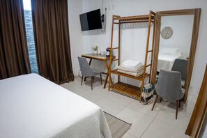 Ein Hotelzimmer mit einem Bett, Schreibtisch, zwei Stühlen, einem Fernseher und einem Holzregal mit gefalteten Handtüchern.