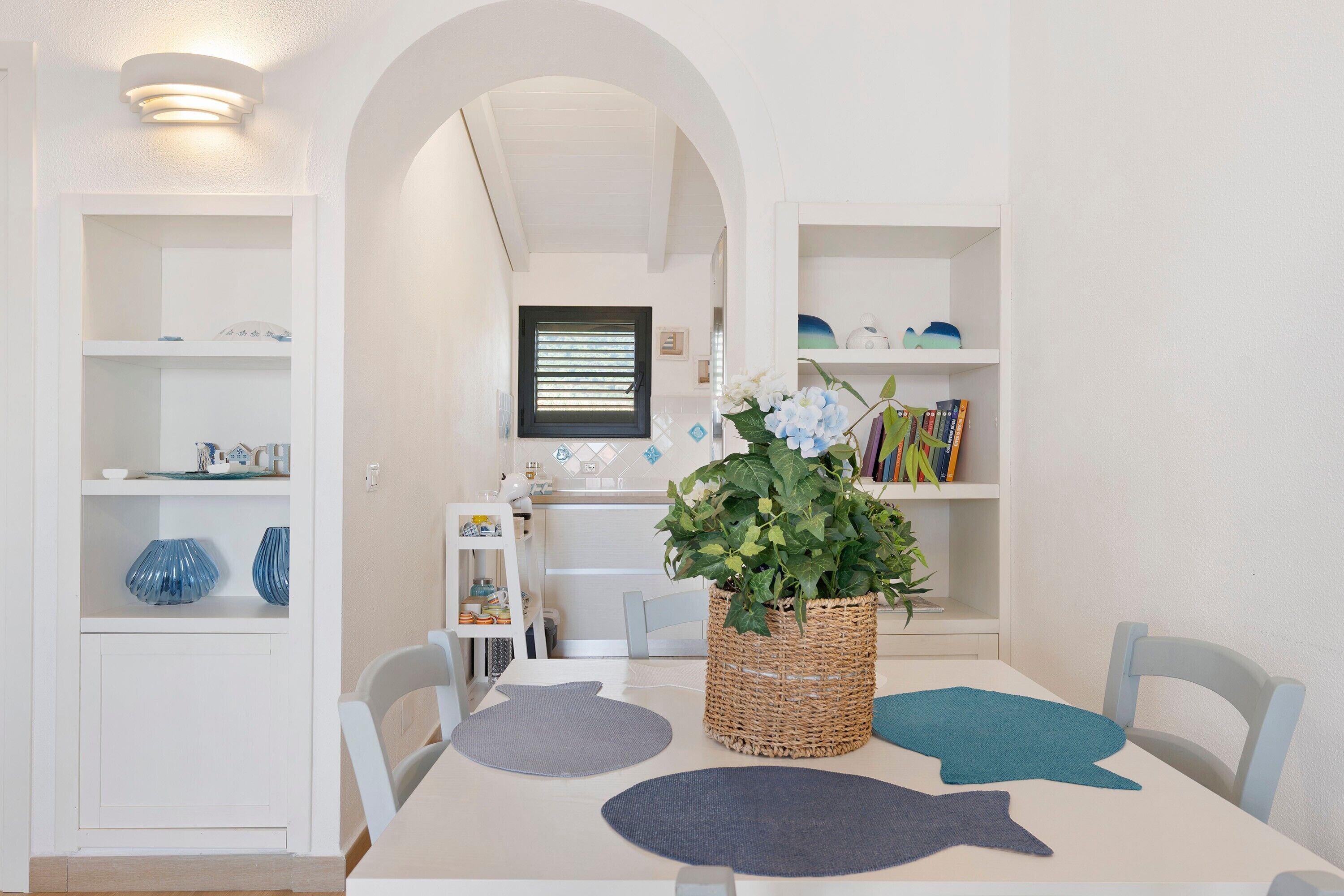 A dining area with a white table, chairs, and a central plant. Shelves with books and decorative items are visible in the background.