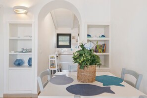 Un coin repas avec une table blanche, des chaises et une plante au centre. On aperçoit, à l’arrière-plan, des étagères remplies de livres et d’objets décoratifs.