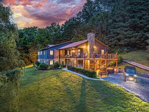 A two-story log cabin with a stone chimney, a deck with outdoor seating, and a driveway leading to a vehicle. - Sleeps 18 · 7BR Estate on 6 Acres Near Bryson City — Creek, Hot Tub, Game Room (Bryson City)