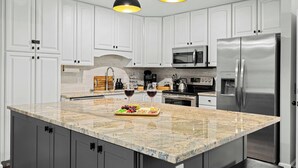 Una cocina con armarios blancos, encimera de granito, electrodomésticos de acero inoxidable e iluminación colgante.