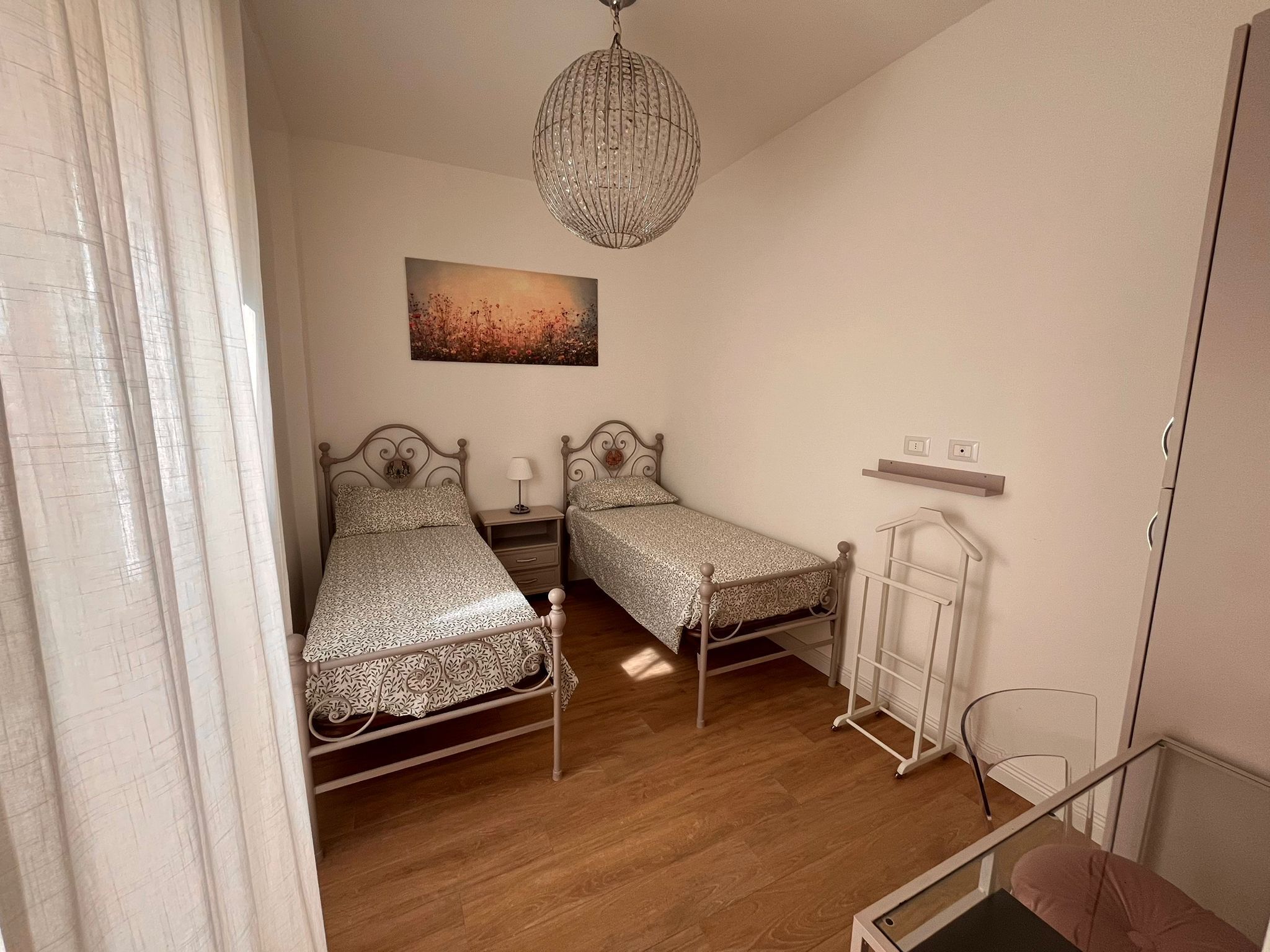 A bedroom with two beds, a chandelier, a painting, and a white chair.