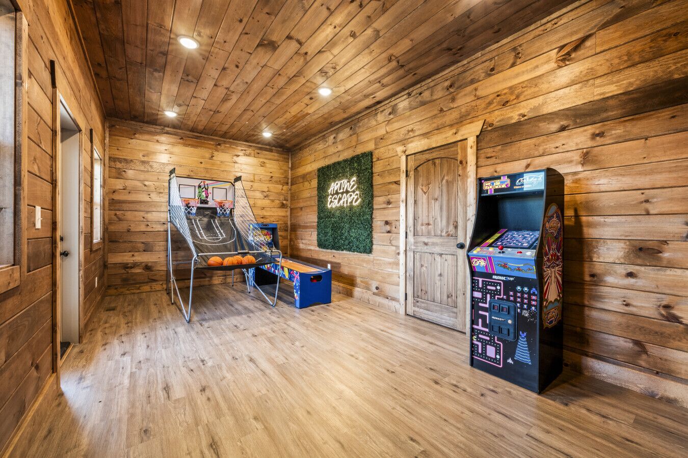 A wooden room with a basketball hoop, arcade game, and a neon sign.