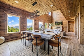 A rustic cabin interior with a large dining table, four chairs, a fireplace, and large windows offering a mountain view. - New! Views, Sauna Spa Hottub Cantina, Dogs (Gatlinburg)