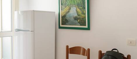 A room with a white table, two wooden chairs, a black backpack on one chair, a white refrigerator, and a framed painting on the wall.