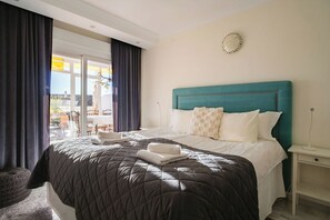 Ein Hotelzimmer mit einem großen Bett, einem türkisfarbenen Kopfteil, einem weißen Nachttisch und Blick ins Freie.