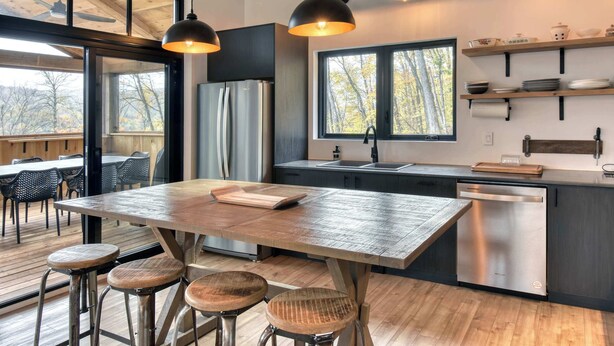 A modern kitchen with a wooden island, bar stools, a dishwasher, and a window with a view of trees.