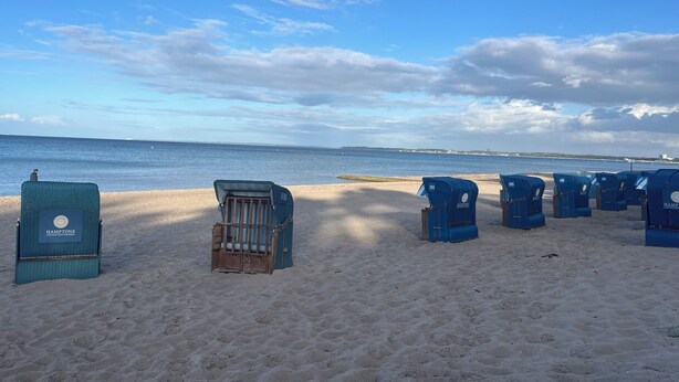Una playa con varias sillas azules para la playa y una silla de madera, arena y un cielo azul despejado.