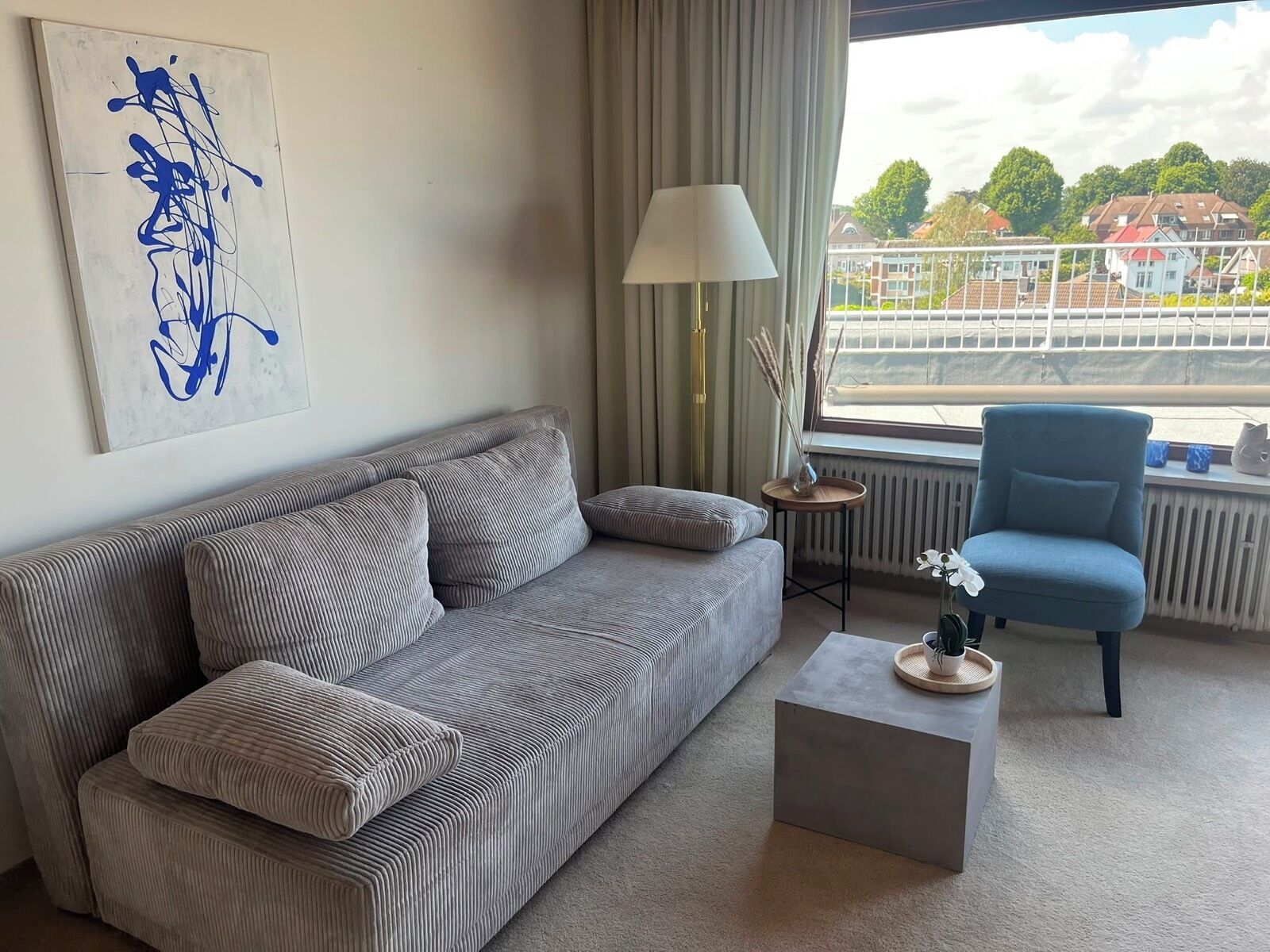 A living room with a sofa, a chair, a lamp, a small table, and a view of buildings through a window.