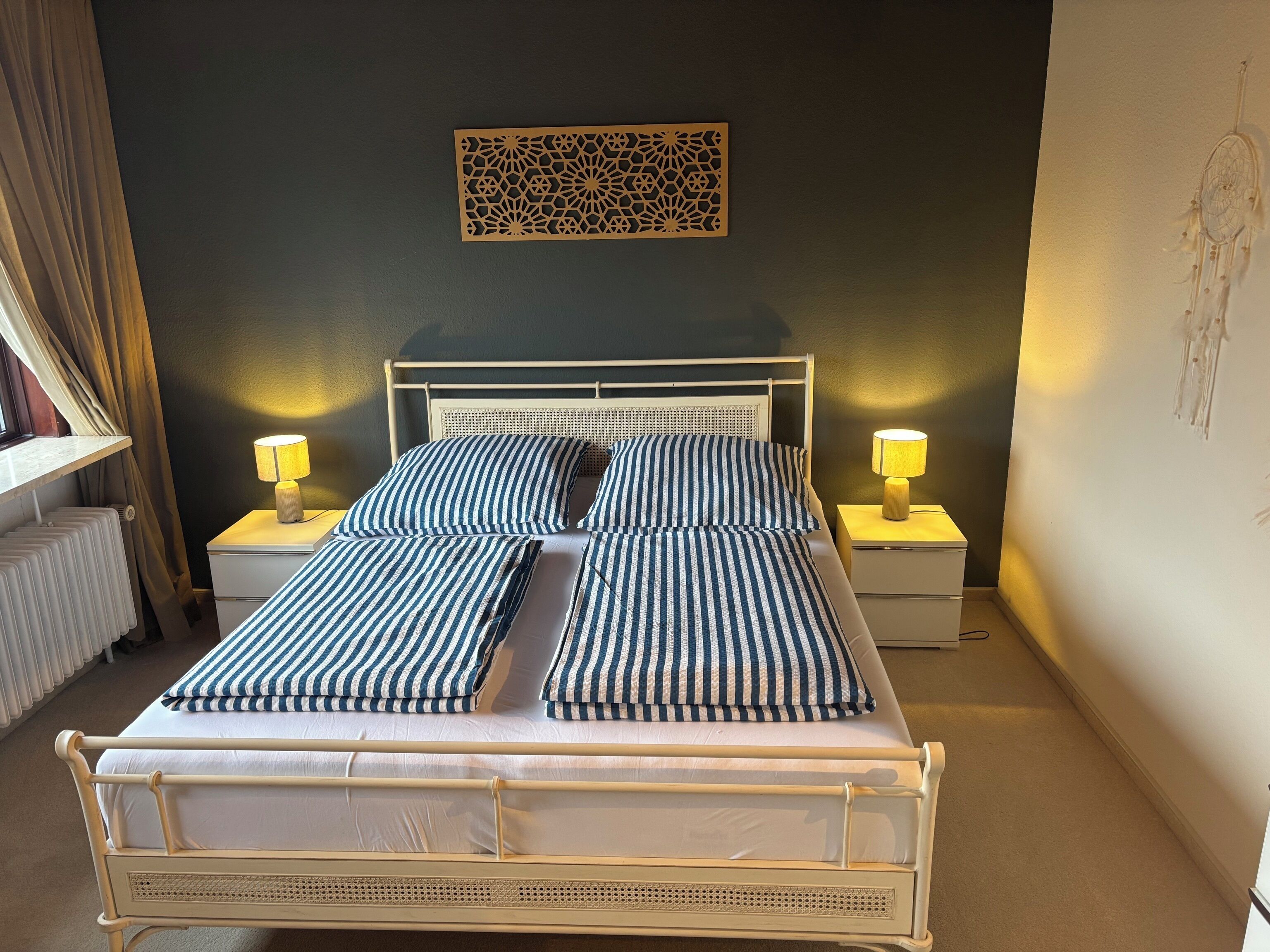 Een bed met gestreept beddengoed, twee bedlampen, een decoratief wandobject en een radiator.
