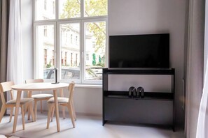 A modern room with a large window, a flat-screen TV on a black stand, a round table with chairs, and a white floor. - Artisan Inspired · Franz Joseph Boutique Studio (Vienna)