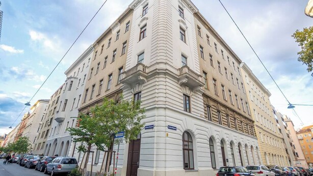 Un edificio de varios pisos con estilo arquitectónico clásico, que presenta una entrada en esquina, una puerta de acceso visible y un letrero que indica un negocio o servicio en la planta baja.