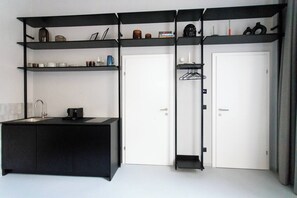 Ein kleines Zimmer mit einer schwarzen Küchenzeile, einer weißen Tür und einem schwarzen Regalelement mit diversen Gegenständen.