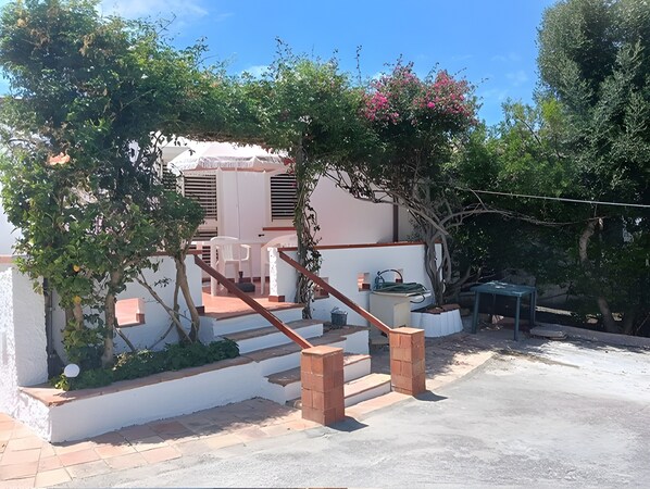 Il s’agit d’une propriété résidentielle dotée d’un escalier menant à un patio couvert, lequel est entouré de verdure et bordé d’un bâtiment blanc.