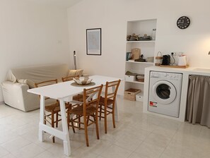 Un espacio habitable compacto con mesa de comedor, sillas, sofá, lavadora y zona de cocina con estantes y un reloj.