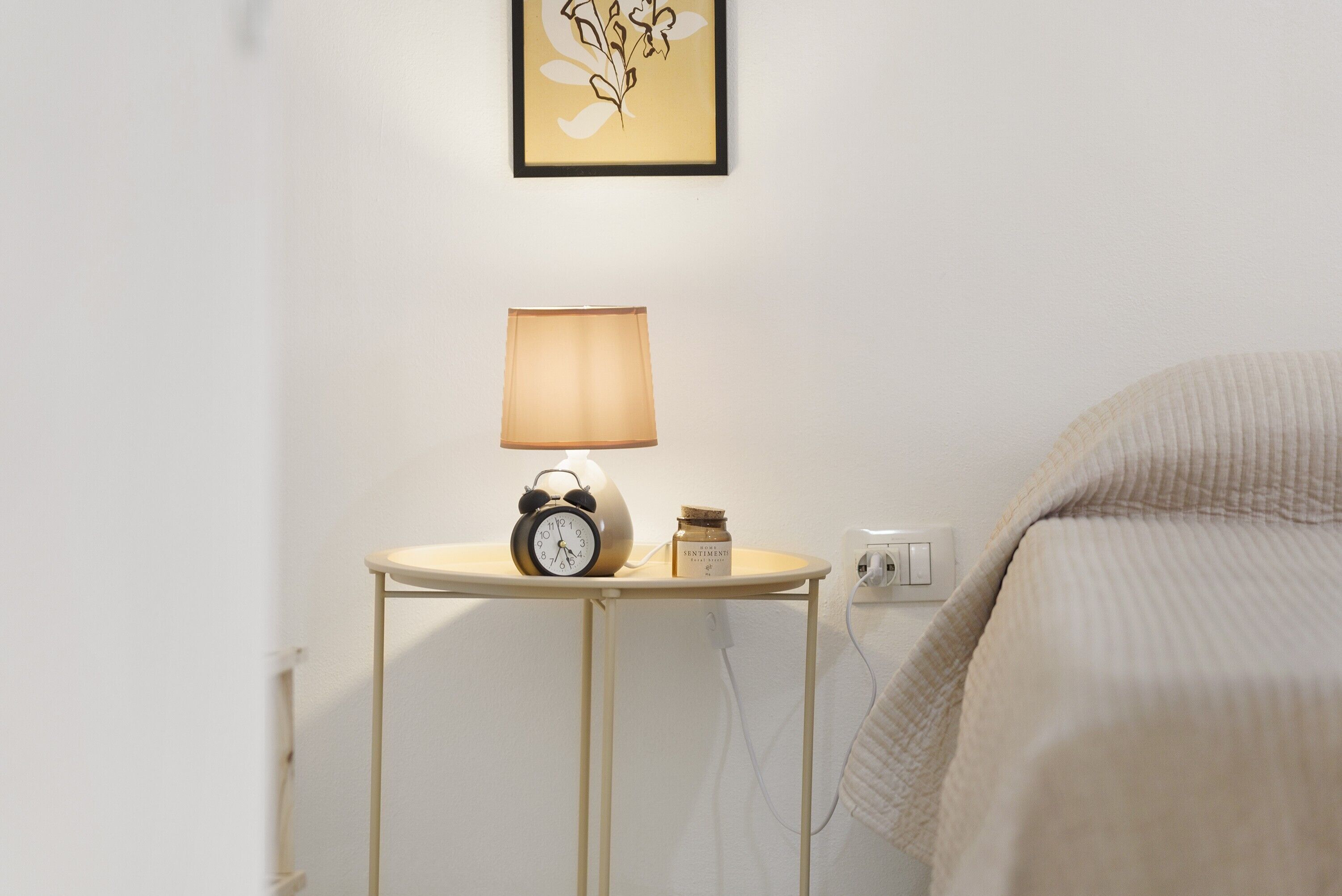 A bedside table with a lamp, an alarm clock, and a small container, likely a candle or jar, against a plain wall with a framed picture above.