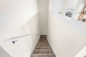 Eine Teppichstufen Treppe mit Handlauf, weißen Wänden und einem Fenster mit Blick auf ein Schlafzimmer.