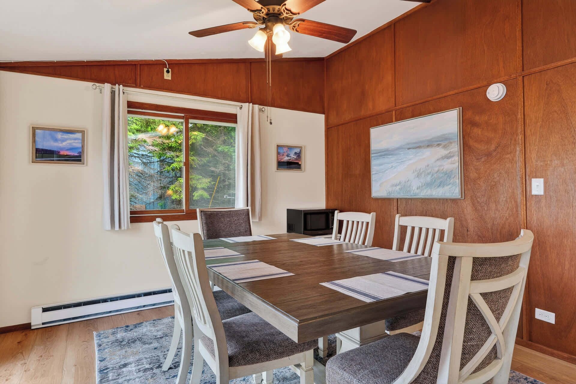 Un coin repas avec une table en bois, des chaises blanches, un ventilateur de plafond et une vue sur les arbres à travers de grandes fenêtres.