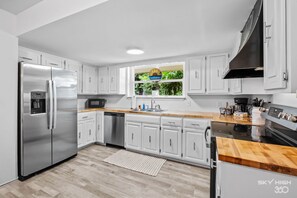A modern kitchen with white cabinets, stainless steel appliances, a wooden countertop, and a window with a view of greenery.