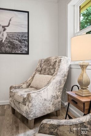 Ein Stuhl mit abgenutztem Blumenmuster, ein gerahmtes Bild an der Wand, eine Lampe auf einem Holztisch und ein Fenster mit Blick auf Grünflächen.