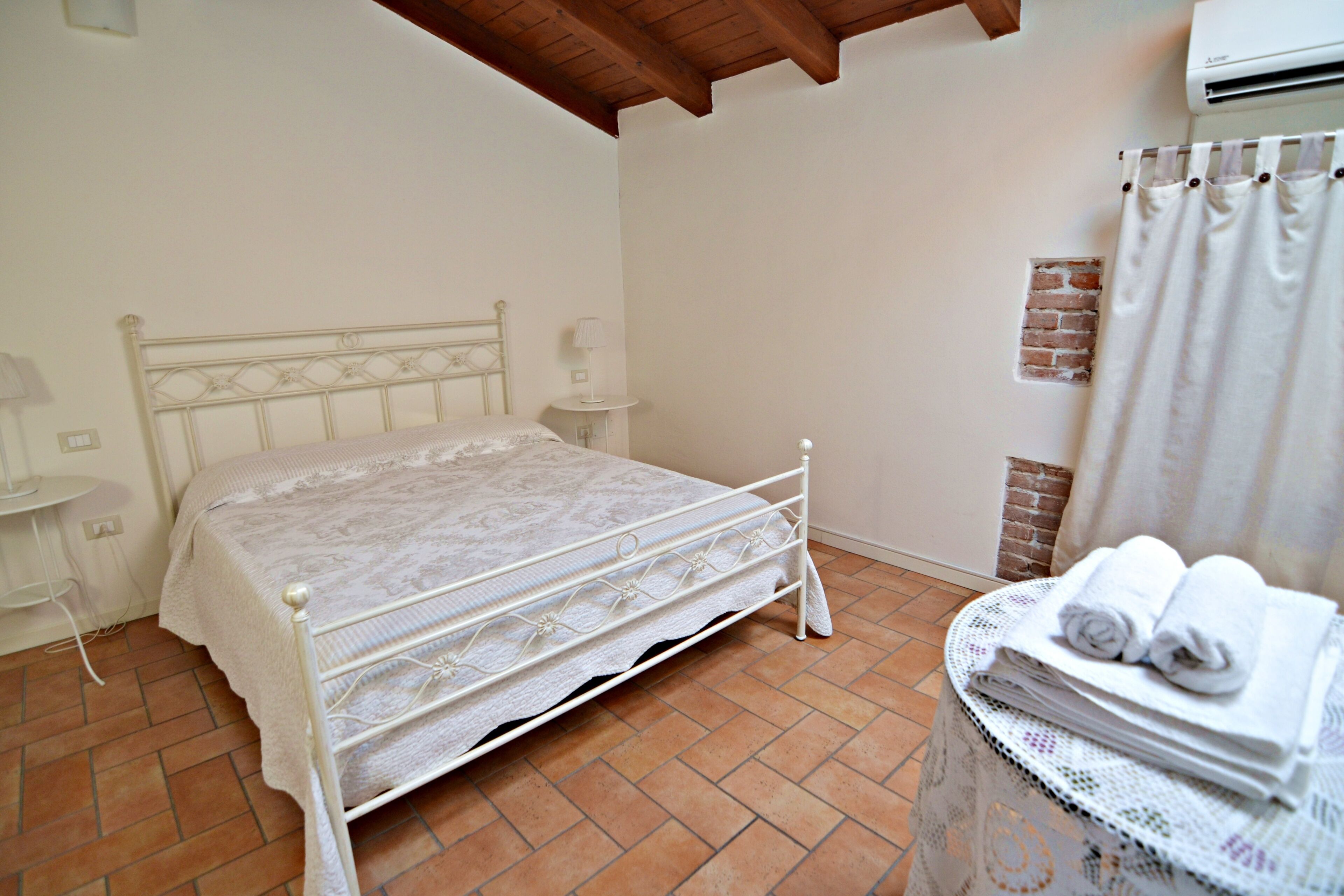 Une chambre avec un lit en métal, une petite table de chevet, une lampe, un climatiseur fixé au mur, un rideau et une table avec des serviettes pliées.