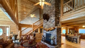 A log cabin interior with a stone fireplace, wooden staircase, and a kitchen area. - Red Rock Lodge — Escape, Relax, and Make Lasting Memories! (Fruitland)
