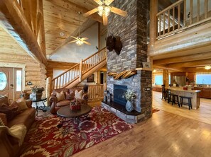A log cabin interior with a stone fireplace, wooden staircase, and a kitchen area. - Red Rock Lodge — Escape, Relax, and Make Lasting Memories! (Fruitland)
