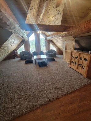 A cozy attic room with a sloped ceiling, a large window, a wooden cabinet with animal carvings, and a television. - Red Rock Lodge — Escape, Relax, and Make Lasting Memories! (Fruitland)