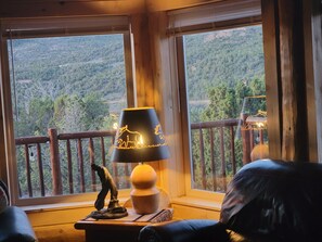 Una stanza con vista su un paesaggio boscoso attraverso ampie finestre, una ringhiera in legno e una lampada con un piedistallo decorativo.