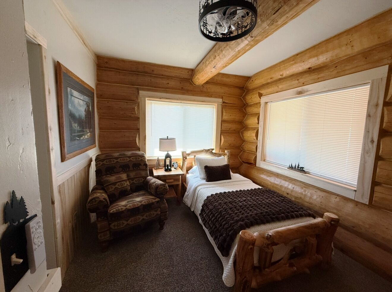A log cabin room with a bed, a chair, a nightstand, and a window with blinds.
