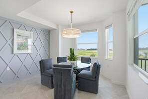 A dining area with a round table, four grey chairs, a hanging light fixture, a framed picture, and large windows with a view of the outdoors. - Golf & Lake View Condo, Resort Pool,  Babcock Ranch (Punta Gorda)