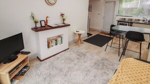 A modern living room with a flat-screen TV, a fireplace, a bookshelf, a dining area with a table and chairs, and a kitchen visible through an open door. - Juniper Hayloft (Ambleside)
