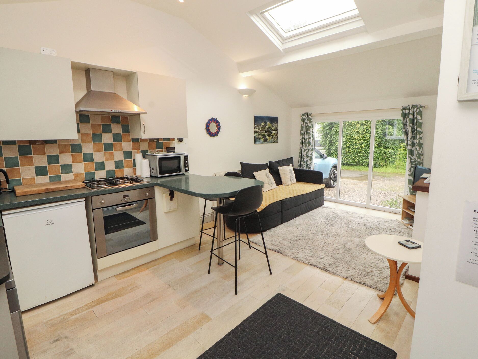 A compact kitchen with a microwave, stove, and sink, a sofa with a view to the outside, and a small dining area.
