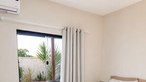 A bedroom with a bed, white curtains, a window with a view of greenery, and an air conditioning unit. - Porto Fino Residential Complex, 5 minutes from the city center. (Porto Seguro)