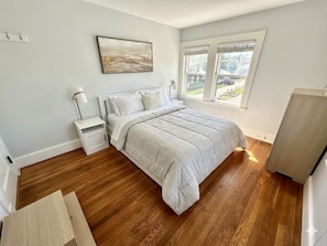 Ein Schlafzimmer mit einem Bett, Nachttischen, einer Lampe, einem Fenster mit Jalousien und Blick ins Freie.