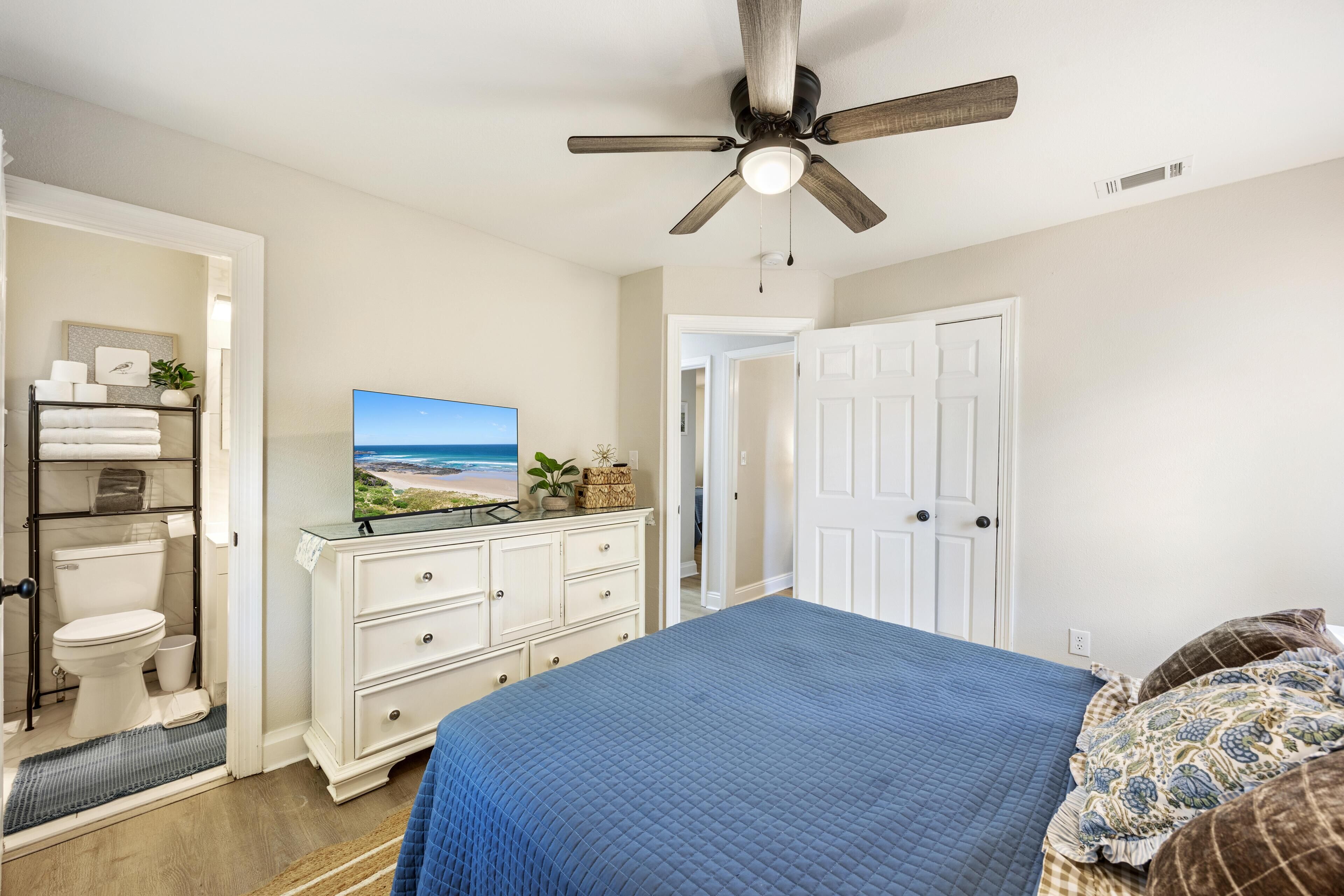 A bedroom with a bed, a ceiling fan, a television, a dresser, a door, and a view of the beach through the window.