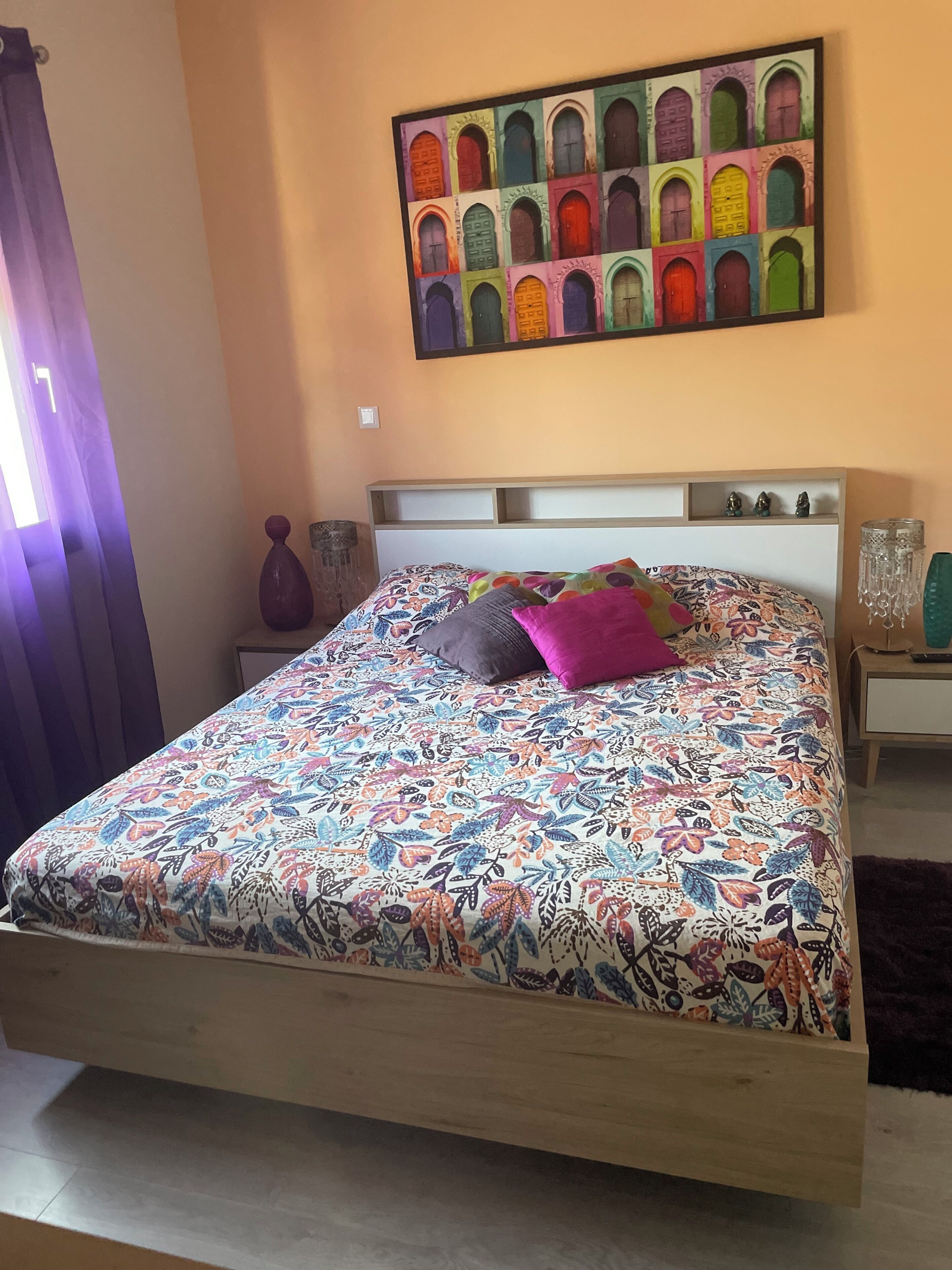 A bedroom with a bed, a nightstand, a wall-mounted painting, and a window with curtains.