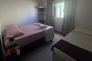 A bedroom with two beds, a bedside table with a lamp, a wardrobe, and a window with curtains.