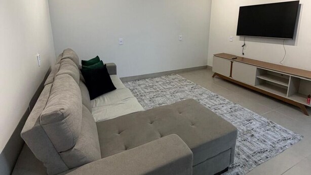 A living room with a sofa, ottoman, and a flat-screen TV mounted on the wall. - Cozy house with 2 air-conditioned bedrooms. (Nova Mutum)