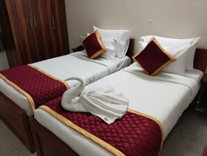 Two beds with white linens and red accents, a wooden wardrobe, and a white towel on one bed.