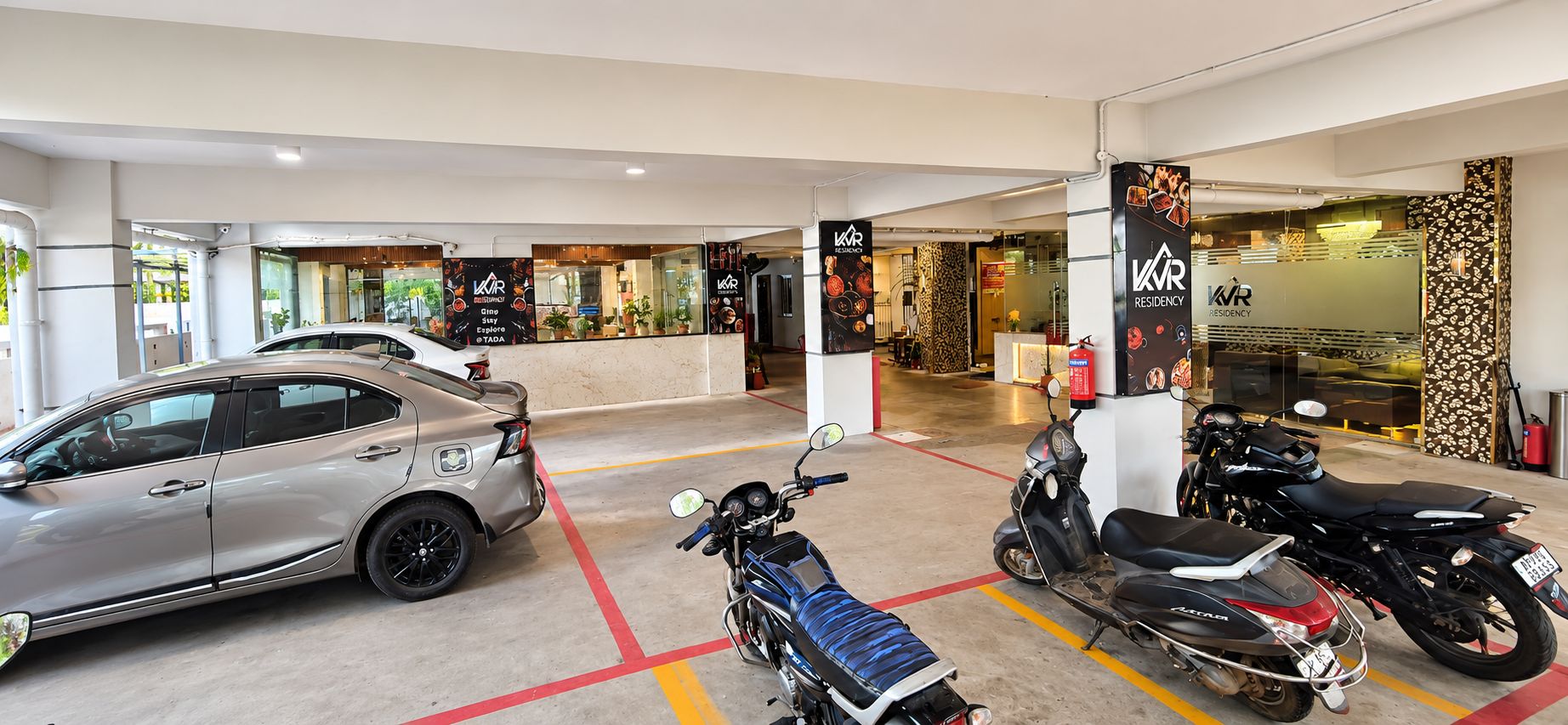 A parking area with a car and two scooters, a reception desk, and a restaurant with a sign for VVR Residency.