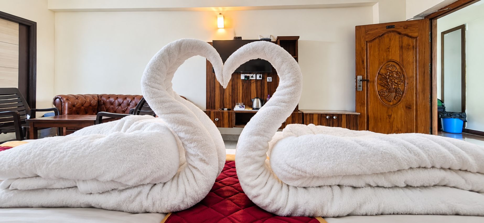 Una camera d'albergo con un letto, una coperta rossa, un divano in pelle marrone, un armadietto in legno e una porta in legno con motivo floreale.