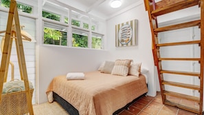A bedroom with a bed, a wooden ladder, a wicker basket, and a window with a view of greenery.