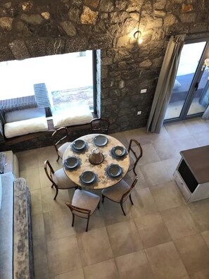A stone-walled dining area with a round table set for six, surrounded by chairs, and large windows offering a view. - House Sleeps 6 With Pool and Parking (Ayia Napa)