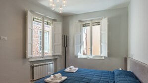 A bedroom with a blue bedspread, two open windows, a radiator, and a black floor lamp.
