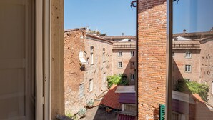 A view through a window of a narrow alleyway with brick buildings, a red-tiled roof, and some greenery.