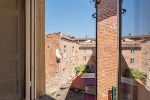 Vistall a través d'una finestra d'un carreró estret, amb edificis de maó, un teulat de teules vermelles i vegetació.