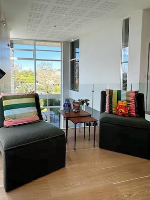 Ein modernes Wohnzimmer mit einer Couch, einem Sessel, einem kleinen Tisch und großen Fenstern.