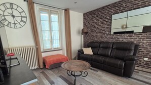 A living room with a brick wall, a leather sofa, a coffee table, a clock, and a window with curtains. - Large family home in the heart of the village (Sainte-Cécile-les-Vignes)